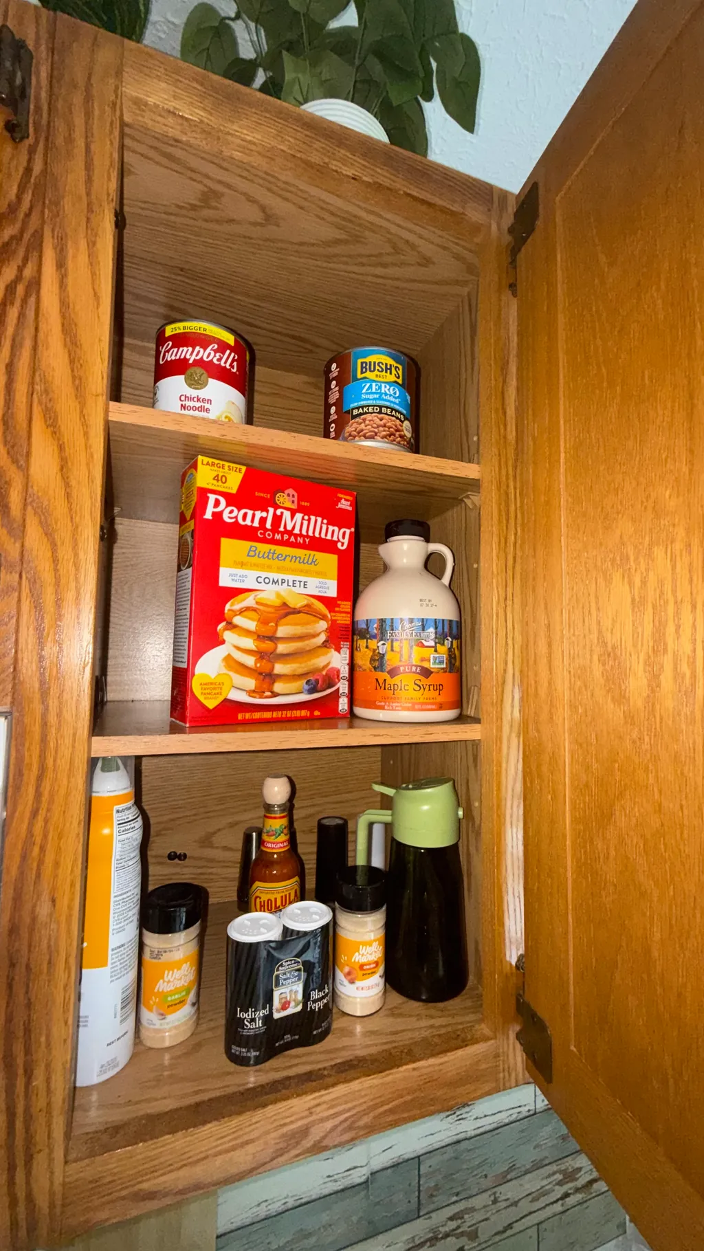 A wooden cabinet with open doors, shelves holding pancake mix, maple syrup, and various condiments.